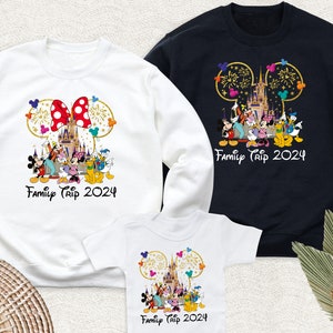 May include: Three white shirts with a Mickey Mouse ear design and a picture of Cinderella's castle with fireworks. The shirts have the text 'Family Trip 2024' printed on them. The shirts are in different sizes: a long-sleeved adult shirt, a short-sleeved adult shirt, and a baby onesie.