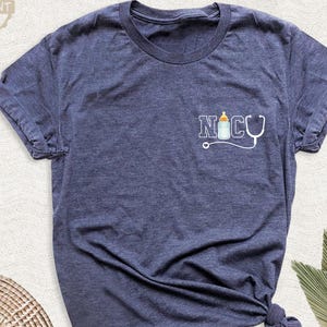 May include: A navy blue t-shirt with a small graphic on the front that says "NICU" with a baby bottle and stethoscope. The shirt has short sleeves and a round neckline. The words "Best Moment Tees" are in a circle at the top left.