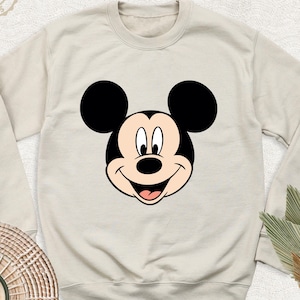 May include: A beige sweatshirt with a black and white Mickey Mouse face graphic on the front.