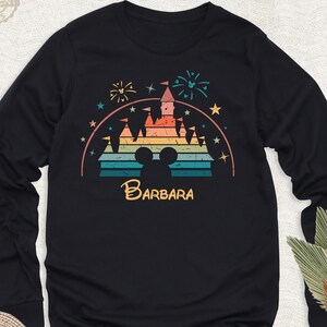 May include: A black long-sleeved shirt with a retro-style illustration of a castle with a rainbow color scheme. The castle is surrounded by stars and fireworks. The text 'BARBARA' is printed below the castle.