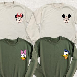 May include: Four crewneck sweatshirts in beige and olive green. Each sweatshirt features a cartoon character face on the chest: Minnie Mouse, Mickey Mouse, Daisy Duck, and Donald Duck. The sweatshirts are long-sleeved and made of a soft material.