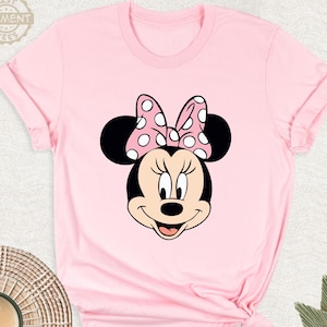 Puede incluir: Camiseta rosa con un dise&ntilde;o de Minnie Mouse. El dise&ntilde;o muestra la cara de Minnie, con orejas negras y un lazo rosa con lunares blancos. La camiseta tiene mangas cortas y cuello redondo.
