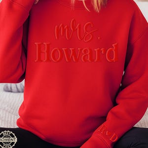 May include: A red sweatshirt with the words "mrs. Howard" in a raised, tone-on-tone design across the chest. The sleeve features the initials "R & D" in a similar style. The sweatshirt is a solid, vibrant red.