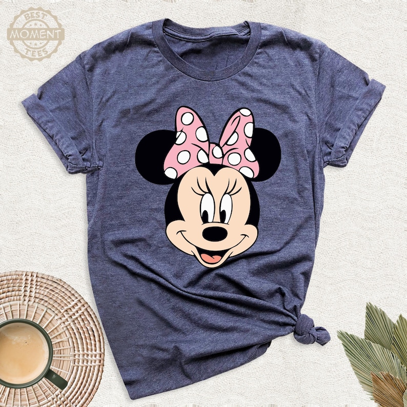 Puede incluir: Una camiseta azul marino con un gr&aacute;fico de Minnie Mouse. El gr&aacute;fico muestra la cara de Minnie con un lazo rosa con lunares blancos. La camiseta tiene mangas cortas y cuello redondo. El texto "BEST MOMENT TEES" est&aacute; en la esquina superior izquierda.