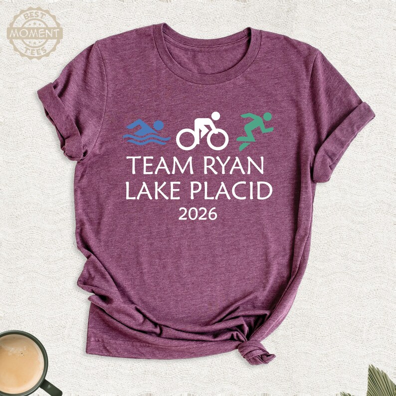 Pu&ograve; includere: T-shirt bordeaux con la scritta bianca "TEAM RYAN LAKE PLACID 2026". Sopra il testo, icone grafiche blu, bianche e verdi che rappresentano nuoto, ciclismo e corsa. La maglietta ha maniche corte e scollo tondo.