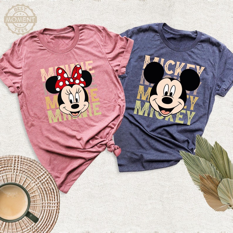 May include: Two t-shirts, one pink and one blue, with a Mickey Mouse and Minnie Mouse design. The pink t-shirt has the text 'Minnie' repeated three times in a vintage style font. The blue t-shirt has the text 'Mickey' repeated three times in a vintage style font.