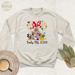 Puede incluir: Una sudadera de color beige claro con un gr&aacute;fico de Mickey Mouse y sus amigos frente al Castillo de Cenicienta. El gr&aacute;fico tambi&eacute;n incluye un lazo rojo con lunares blancos y el texto "Family Trip 2024".