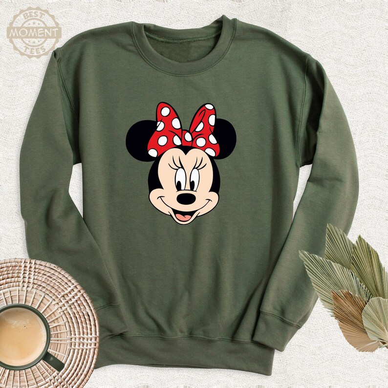 Peut inclure: Un sweat-shirt vert avec une image de dessin anim&eacute; de Minnie Mouse. Minnie Mouse porte un n&oelig;ud rouge &agrave; pois blancs.