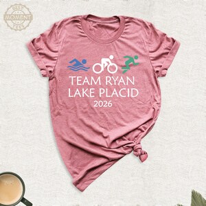 Pu&ograve; includere: T-shirt rosa polveroso con la scritta bianca "TEAM RYAN LAKE PLACID 2026". Sopra il testo ci sono icone grafiche blu, bianche e verdi di nuoto, ciclismo e corsa. La maglietta &egrave; realizzata in materiale morbido.