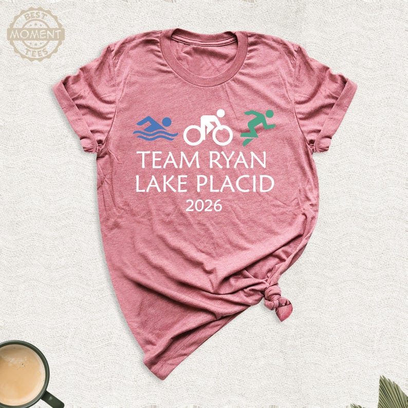Pu&ograve; includere: T-shirt rosa polveroso con la scritta bianca "TEAM RYAN LAKE PLACID 2026". Sopra il testo ci sono icone grafiche blu, bianche e verdi di nuoto, ciclismo e corsa. La maglietta &egrave; realizzata in materiale morbido.