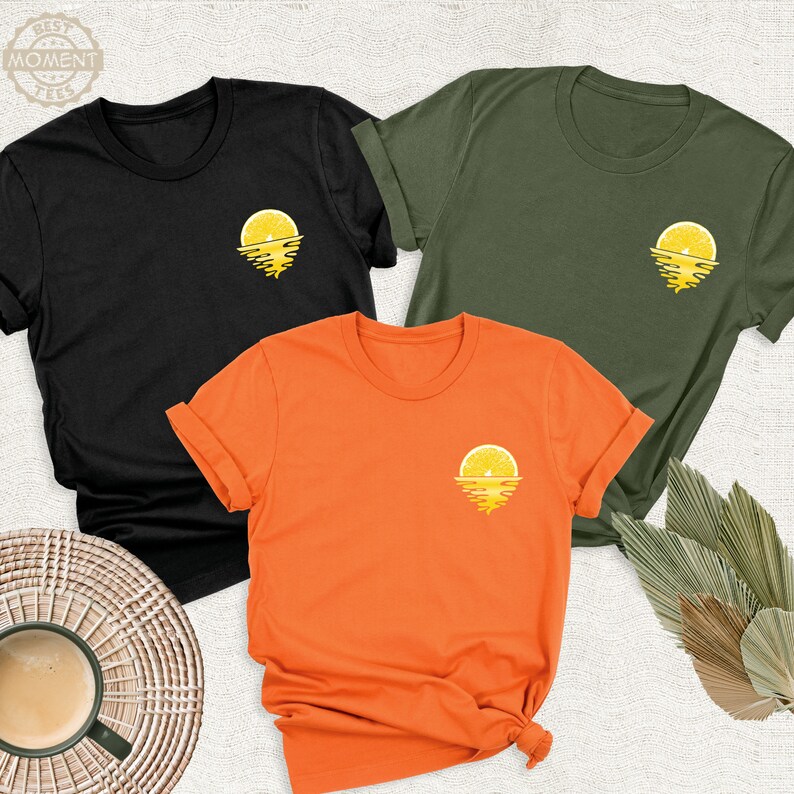 May include: Three t-shirts in black, olive green, and orange with a lemon slice sunset graphic on the front.