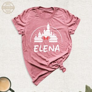 Puede incluir: Camiseta rosa polvoriento con un gr&aacute;fico blanco de un castillo, un lazo rojo y el nombre "ELENA". El dise&ntilde;o est&aacute; impreso en la parte delantera. La camiseta tiene cuello redondo y mangas cortas. El material parece suave.