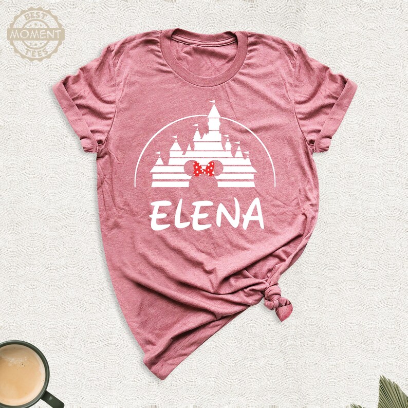 Puede incluir: Camiseta rosa polvoriento con un gr&aacute;fico blanco de un castillo, un lazo rojo y el nombre "ELENA". El dise&ntilde;o est&aacute; impreso en la parte delantera. La camiseta tiene cuello redondo y mangas cortas. El material parece suave.