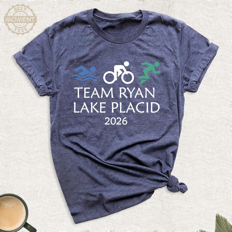 Pu&ograve; includere: T-shirt blu navy con la scritta bianca "TEAM RYAN LAKE PLACID 2026". Il design include figure di nuoto blu, una bicicletta bianca e una figura di corridore verde, che rappresenta un triathlon.