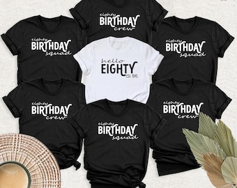 May include: A collection of six short-sleeved t-shirts. Five are black with white text reading "eighty BIRTHDAY squad" or "eighty BIRTHDAY crew". One central white t-shirt features black text reading "hello EIGHTY est. 1945".