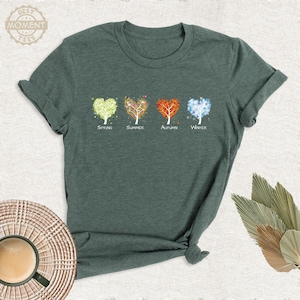 May include: A dark green short-sleeved t-shirt featuring a graphic of four trees shaped like hearts, representing the four seasons. Below each tree, the words Spring, Summer, Autumn, and Winter are printed in a simple white font.