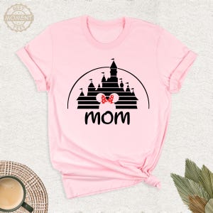 Puede incluir: Camiseta rosa claro con una silueta negra de castillo y un lazo de lunares rojos y blancos. La palabra "mam&aacute;" est&aacute; impresa en negro debajo del castillo. La camiseta est&aacute; doblada en la parte inferior.