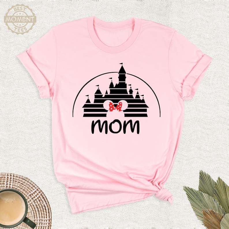 Puede incluir: Camiseta rosa claro con una silueta negra de castillo y un lazo de lunares rojos y blancos. La palabra "mam&aacute;" est&aacute; impresa en negro debajo del castillo. La camiseta est&aacute; doblada en la parte inferior.