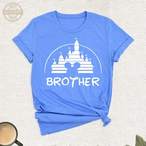 Puede incluir: Camiseta azul claro con un gr&aacute;fico blanco de un castillo, orejas de Mickey Mouse y la palabra "BROTHER". La camiseta tiene mangas enrolladas y un dobladillo anudado.