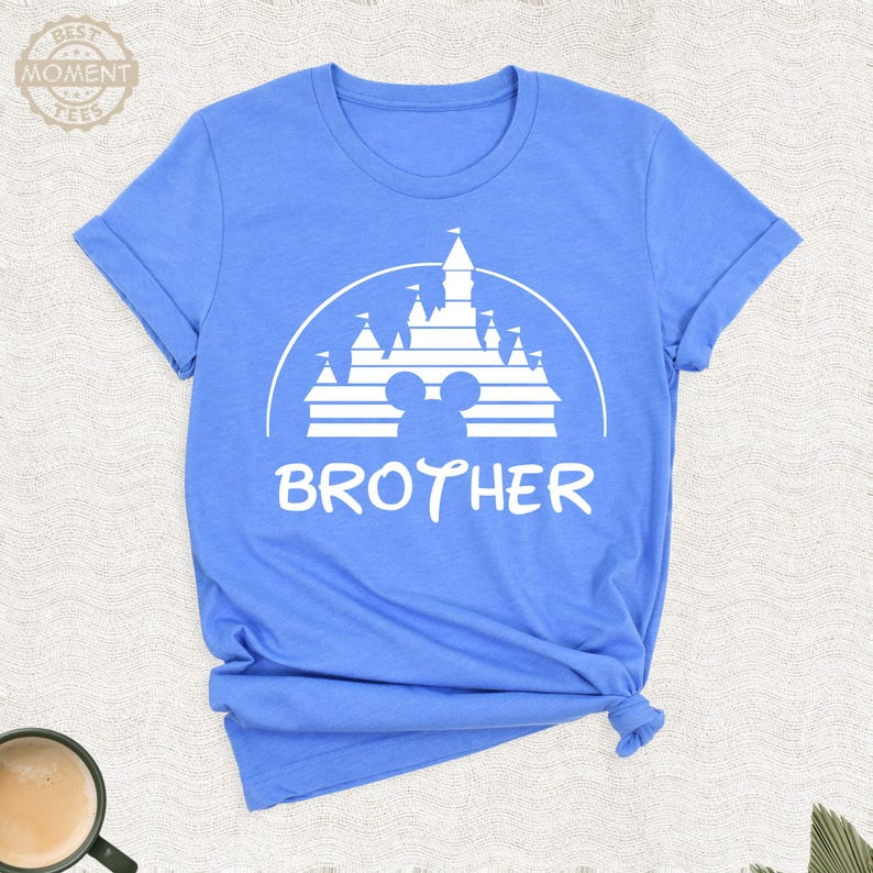 Puede incluir: Camiseta azul claro con un gr&aacute;fico blanco de un castillo, orejas de Mickey Mouse y la palabra "BROTHER". La camiseta tiene mangas enrolladas y un dobladillo anudado.