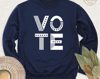 May include: A navy blue crewneck sweatshirt featuring the word VOTE in large white letters. The letter V is composed of vertical stripes and stars, the O is a circle with stars, the T has a row of stars, and the E has horizontal stripes.