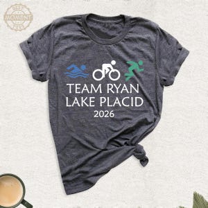 Pu&ograve; includere: T-shirt grigio scuro con la scritta "TEAM RYAN LAKE PLACID 2026" e grafica a tema triathlon in blu, bianco e verde. Il design include un nuotatore, un ciclista e un corridore.