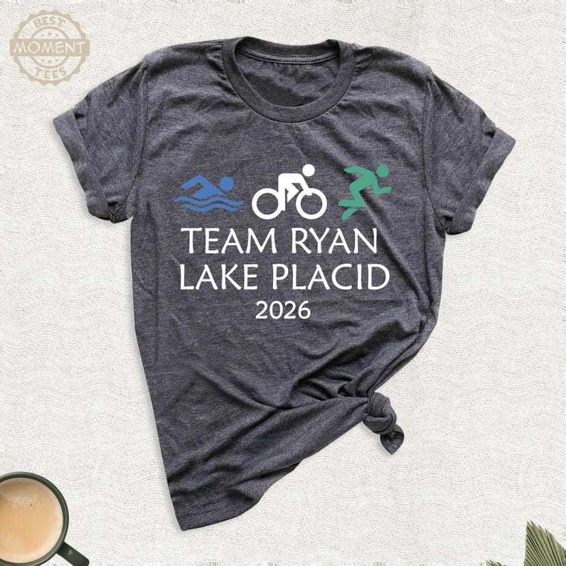 Pu&ograve; includere: T-shirt grigio scuro con la scritta "TEAM RYAN LAKE PLACID 2026" e grafica a tema triathlon in blu, bianco e verde. Il design include un nuotatore, un ciclista e un corridore.