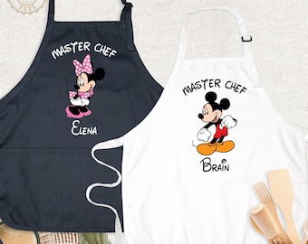 May include: A black apron featuring Minnie Mouse and the text "MASTER CHEF ELENA" and a white apron featuring Mickey Mouse and the text "MASTER CHEF BRAIN". Both aprons have adjustable neck straps and front pockets for kitchen use.