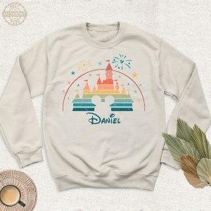 May include: A light beige sweatshirt with a retro-style illustration of a castle with a rainbow color scheme. The castle is surrounded by stars and fireworks. The text "Daniel" is printed below the castle.