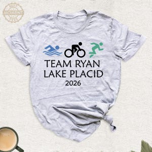 Pu&ograve; includere: T-shirt grigio chiaro con la scritta "TEAM RYAN LAKE PLACID 2026". Il design include onde blu di nuoto, un ciclista nero e un corridore verde, che rappresentano un triathlon.