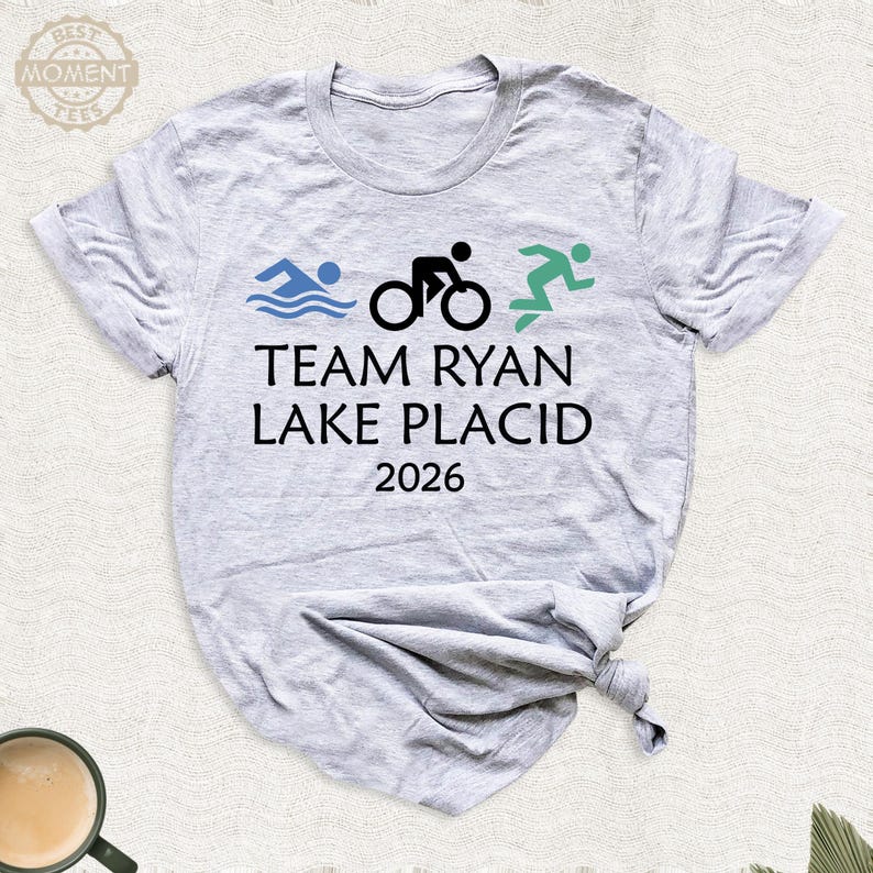 Pu&ograve; includere: T-shirt grigio chiaro con la scritta "TEAM RYAN LAKE PLACID 2026". Il design include onde blu di nuoto, un ciclista nero e un corridore verde, che rappresentano un triathlon.