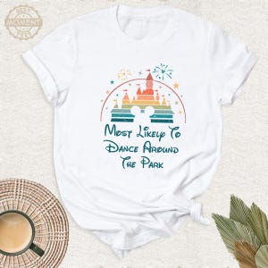 May include: White t-shirt featuring a colorful graphic of a castle, fireworks, and Mickey Mouse ears. The text reads "Most Likely To Dance Around The Park" in a whimsical font. The shirt is made of soft material.