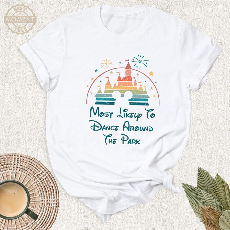 May include: White t-shirt featuring a colorful graphic of a castle, fireworks, and Mickey Mouse ears. The text reads "Most Likely To Dance Around The Park" in a whimsical font. The shirt is made of soft material.