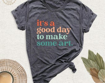 May include: A dark heather gray short-sleeve t-shirt featuring the text "it's a good day to make some art" printed in orange, light peach, and teal. The shirt has a crew neckline and a knotted hem detail on the lower right side of the garment.