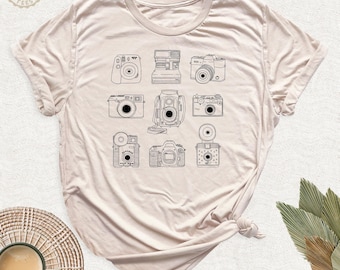 May include: A light beige short-sleeve t-shirt featuring a black line art graphic print of nine different vintage cameras arranged in a grid. The design includes various camera styles, including box, rangefinder, and instant film models.