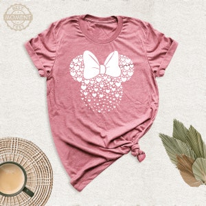 Puede incluir: Camiseta rosa polvoriento con un dise&ntilde;o de Minnie Mouse blanco hecho de corazones y un lazo. El texto "BEST MOMENT TEES" est&aacute; en un logotipo circular en la parte superior izquierda. La camiseta est&aacute; anudada en la parte inferior.