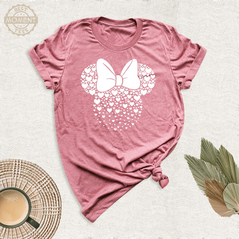 Puede incluir: Camiseta rosa polvoriento con un dise&ntilde;o de Minnie Mouse blanco hecho de corazones y un lazo. El texto "BEST MOMENT TEES" est&aacute; en un logotipo circular en la parte superior izquierda. La camiseta est&aacute; anudada en la parte inferior.