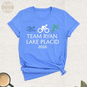 Pu&ograve; includere: Maglietta azzurra con la scritta bianca "TEAM RYAN LAKE PLACID 2026". Sopra, icone di nuoto, ciclismo e corsa. La maglietta ha maniche arrotolate e un nodo in basso. Perfetta per eventi sportivi.