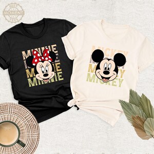 May include: Two t-shirts with a retro design featuring Mickey Mouse and Minnie Mouse. The Mickey Mouse shirt is beige with a brown, yellow, and green color scheme. The Minnie Mouse shirt is black with a red, yellow, and green color scheme.