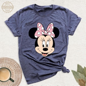 Puede incluir: Una camiseta azul marino con un gr&aacute;fico de Minnie Mouse. El gr&aacute;fico muestra la cara de Minnie con un lazo rosa con lunares blancos. La camiseta tiene mangas cortas y cuello redondo. El texto "BEST MOMENT TEES" est&aacute; en la esquina superior izquierda.