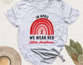 May include: A light gray short-sleeve t-shirt featuring a red rainbow graphic with a central red ribbon. The text reads IN APRIL WE WEAR RED Autism Acceptance in black and red lettering. The shirt has a crew neckline and a knotted hem detail.