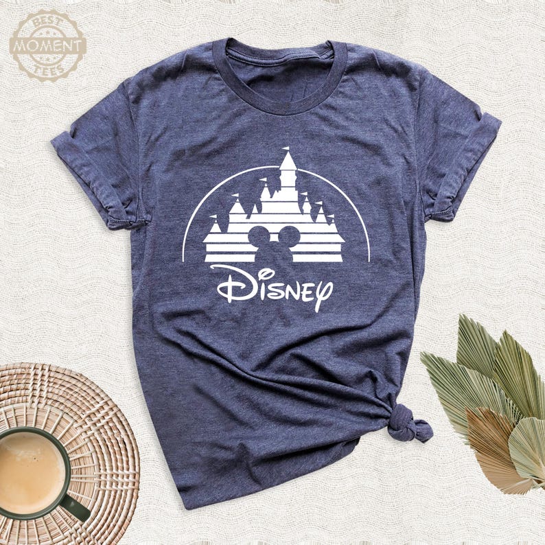 May include: Navy blue t-shirt featuring a white graphic of a castle, Mickey Mouse ears, and the word "Disney". The shirt has short sleeves and a crew neck. The shirt is made of a soft, comfortable material.