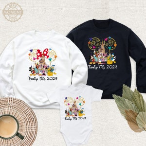 Puede incluir: Camisas blancas, negras y azul beb&eacute; con un dise&ntilde;o de orejas de Mickey Mouse que presenta el castillo de Disney, fuegos artificiales y personajes de Mickey Mouse. Las camisas tienen el texto "Family Trip 2024".