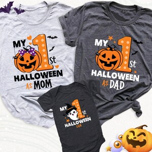 K&ouml;nnte beinhalten: Drei Halloween-Shirts. Zwei T-Shirts f&uuml;r Erwachsene, eines grau und eines dunkelgrau, zeigen eine K&uuml;rbisgrafik und den Text "My 1st Halloween As Mom" und "My 1st Halloween As Dad". Ein dunkelgrauer Baby-Body hat eine Geistergrafik und den Text "My 1st Halloween".