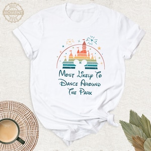 May include: White t-shirt featuring a colorful graphic of a castle, fireworks, and Mickey Mouse ears. The text reads "Most Likely To Dance Around The Park" in a whimsical font. The shirt is made of soft material.
