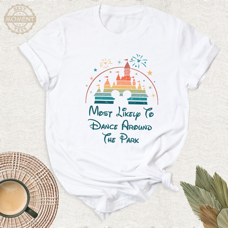 May include: White t-shirt featuring a colorful graphic of a castle, fireworks, and Mickey Mouse ears. The text reads "Most Likely To Dance Around The Park" in a whimsical font. The shirt is made of soft material.