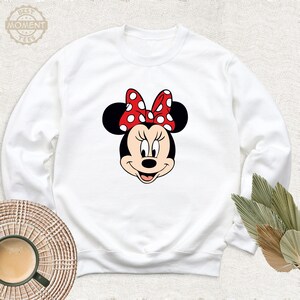 Peut inclure: Sweat-shirt blanc &agrave; manches longues avec une image de dessin anim&eacute; rouge et blanc de Minnie Mouse portant un n&oelig;ud rouge &agrave; pois blancs.