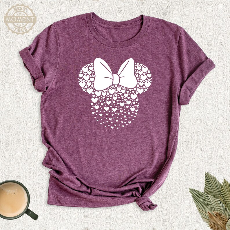 Puede incluir: Una camiseta granate con un dise&ntilde;o de Minnie Mouse blanco. El dise&ntilde;o est&aacute; hecho de corazones y un lazo. El texto "BEST MOMENT TEES" est&aacute; en la esquina superior izquierda. La camiseta est&aacute; doblada en las mangas y anudada en la parte inferior.