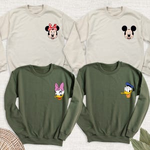 May include: Four crew neck sweatshirts in beige and olive green. Each sweatshirt features a cartoon character face on the chest: Minnie Mouse, Mickey Mouse, Daisy Duck, and Donald Duck. The sweatshirts are long-sleeved and made of a soft material.
