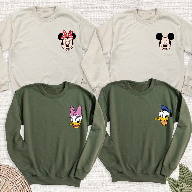 May include: Four crew neck sweatshirts in beige and olive green. Each sweatshirt features a cartoon character face on the chest: Minnie Mouse, Mickey Mouse, Daisy Duck, and Donald Duck. The sweatshirts are long-sleeved and made of a soft material.